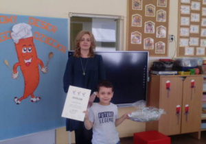 Uśmiechnięty chłopiec, trzymający dyplom i nagrodę na zdjęciu wraz dyrektorem przedszkola.